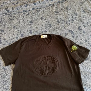 Svart Stone Island t-shirt med emblem - Svart t-shirt från Stone Island i bomull med stort präglat logomärke på bröstet och klassisk patch med kompass på ärmen. Enkel och stilren modell med rund hals och korta ärmar. Perfekt för dig som gillar streetwear och vill sticka ut med en ikonisk detalj.