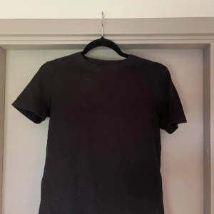 Svart t-shirt från Lyle & Scott Junior - En stilren svart t-shirt från Lyle & Scott Junior. Klassisk rund hals och korta ärmar, perfekt för en enkel och clean look. Tillverkad i mjuk bomull som känns skön mot huden. Passar dig som gillar basic och tidlös stil.