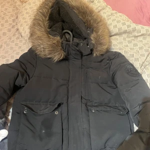 Svart dunjacka med päls på superdry - Säljer en svart dunjacka från Superdry med stor huva och riktigt päls. Jackan har flera fickor med knappar, broderad logga på ärmen och Superdry-brodyr på ryggen. Perfekt för kalla dagar och riktigt snygg streetstil. Dragkedjan har fastnat lite men de går alltid och fixa den är använd max 4 gånger nypris 1600