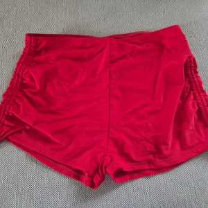 Röda shorts med rynkade sidor - Snygga röda shorts med rynkade detaljer längs sidorna. De är tillverkade i ett stretchigt material som består av 80% polyamid och 20% elastan, vilket gör dem perfekta för träning eller bad. Modellen är kort och sitter tajt mot kroppen. Aldrig använda!