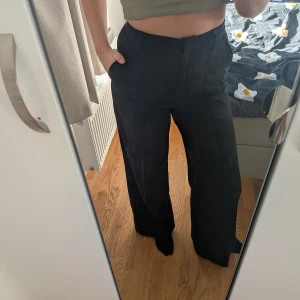 Svarta vida kostymbyxor - Säljer ett par svarta kostymbyxor med hög midja och vida ben. Byxorna har pressveck framtill och sidfickor, vilket ger en clean och stilren look. Perfekta för dig som gillar en relaxed men ändå dressad stil.
