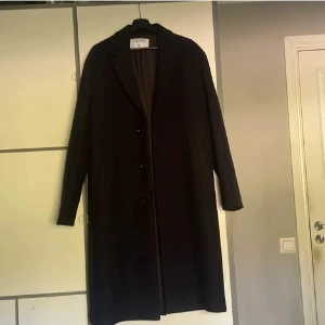 Svart lång kappa från Filippa K - Stilren svart kappa från Filippa K i klassisk rak modell med knappar framtill och krage. Perfekt för dig som gillar minimalistisk och tidlös design. Kappan är tillverkad i ullblandning och har långa ärmar. Passar dig som vill ha en clean och snygg look.