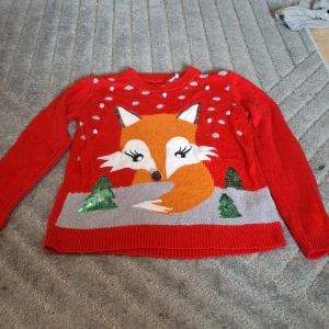 Röd stickad jultröja med rävmotiv Gina Tricot - Supermysig röd stickad tröja från Gina Tricot med ett stort rävmotiv framtill, snöprickar och glittriga paljettgranar. Perfekt för julen med sin färgglada och lekfulla design. Rund halsringning och långa ärmar. Har ett litet hål vid halsen.
