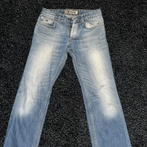 Ljusblå jeans från Tommy Hilfiger - Säljer ett par ljusblå jeans från Tommy Hilfiger med snyggt slitna detaljer och klassisk femficksdesign. Jeansen är raka i modellen och tillverkade i 100% bomull. Perfekta för dig som gillar en avslappnad och tidlös look. Den är äkta jag köpte den för 859kr men kvitto är borta så tyvärr, och den är typ lite baggy straight jeans 