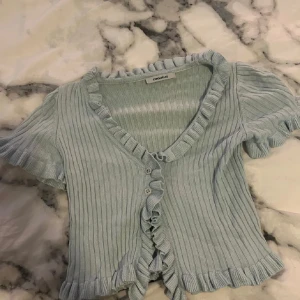 Ljusblå ribbad blus från Chiquelle - Superfin ljusblå blus från Chiquelle med ribbad struktur och volangdetaljer längs v-ringningen, ärmslut och nederkant. Kortärmad och med knappar framtill, perfekt för en söt och trendig look. Passar dig som gillar romantiska vibes och pastellfärger.