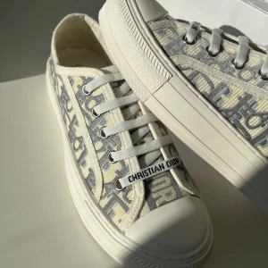 Christian Dior gråvita sneakers - Snygga sneakers från Christian Dior med grått och vitt monogrammönster, platt sula och rund tå. Skorna har snörning med vita skosnören och detaljer med logotypen på tungan. Tillverkade i textil och skinn, med cool stjärndetalj under sulan.