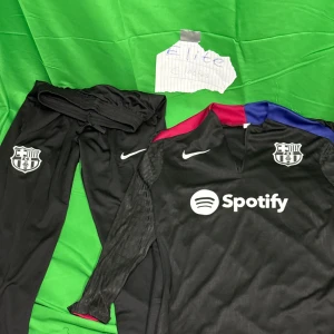 FC Barcelona svart långärmad fotbollströja - Svart långärmad FC Barcelona fotbollströja från Nike med klubbmärke och Spotify-logga på bröstet. Tröjan har detaljer i rött och blått vid axlarna och är tillverkad i ett lätt, sportigt material. Matchande svarta byxor med klubbmärke och Nike-logga ingår.Liknande modell som originalet!