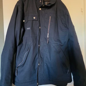 Svart jacka från Depalma Workwear XL - Svart jacka från Depalma Workwear i storlek XL. Jackan har flera fickor med dragkedjor och tryckknappar, hög krage och en clean, stilren look. Perfekt för dig som gillar funktionella detaljer och streetwear-vibe.