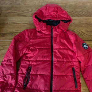 Röd pufferjacka från Everest - Riktigt snygg röd pufferjacka från Everest med avtagbar huva och svarta dragkedjor. Jackan har quiltad design, två fickor med dragkedja och en patch på ärmen. Perfekt för kyliga dagar och enkel att matcha med din stil. Tjock jacka för vinter.
