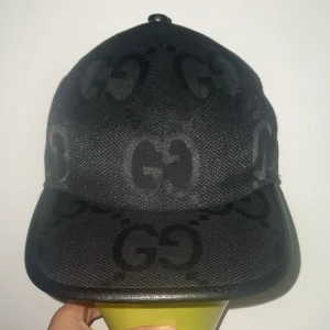 Svart keps från Gucci med GG-mönster - Säljer en svart keps från Gucci med klassiskt GG-mönster över hela kepsen. Kepsen har böjd skärm, justerbar rem baktill och är tillverkad i ett slitstarkt tyg med detaljer i läder. Perfekt för dig som vill ha en stilren och lyxig accessoar.