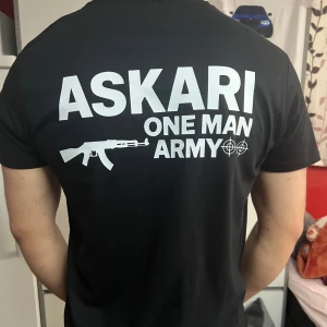 Svart ASKARI t-shirt med tryck - Svart t-shirt från ASKARI med vit text och motiv på både bröstet och ryggen. Trycket har texten 'ASKARI ONE MAN ARMY' tillsammans med ett gevär och måltavlor. Klassisk rund hals och korta ärmar. Skön passform och stilren design.