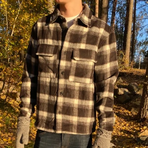 Rutig overshirt i svart och beige - Snygg overshirt i svart, beige och vitt rutigt mönster. Skjortan har klassisk krage, två stora bröstfickor med lock och knäppning framtill. Tillverkad i mjukt bomullstyg, perfekt att slänga över en t-shirt för en avslappnad look. Från H&M, skick 10/10