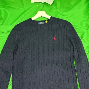 Mörkblå kabelstickad tröja Polo Ralph Lauren - Kabelstickad mörkblå tröja från Polo Ralph Lauren med klassisk rund halsringning och röd broderad logga på bröstet. Långärmad och i mjuk bomull, perfekt för lager-på-lager. Snygg och tidlös stil som funkar året runt.Liknande modell som originalet!