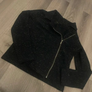 Svart glittrig jacka från Top Secret - Svart jacka från Top Secret med glittriga inslag och sned dragkedja. Jackan har en klassisk krage och är långärmad. Materialet är stickat och ger en mjuk känsla. Perfekt för dig som vill sticka ut lite extra.