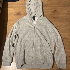 Ljusgrå hoodie med dragkedja H&M - Säljer en ljusgrå hoodie från H&M med hel dragkedja framtill och huva. Tröjan har långa ärmar, ribbade muddar och två fickor på sidorna. Perfekt för chill dagar eller när du vill ha en enkel och clean look.