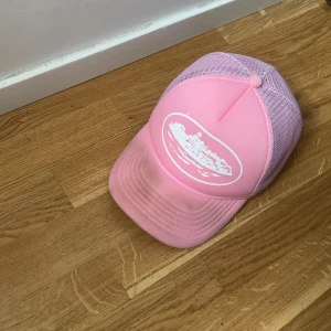 Cortiez keps - Säljer en rosa truckerkeps från Corteiz med vit broderad logga bak och stjärnor på sidan. Framsidan har ett vitt tryck med motiv och kepsen har meshpaneler för extra ventilation. Justerbar snapback och cool streetstil. Går att tvätta. Pris kan diskuteras 