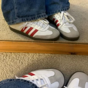 Adidas Samba vita och röda sneakers - Säljer ett par klassiska Adidas Samba sneakers i vitt skinn med röda ränder och detaljer. Skorna har beige mockadetaljer vid tån, gummisula och snörning. Sparsamt använda! 