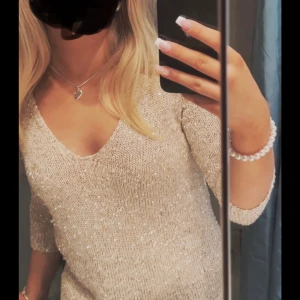 Glittrig beige stickad tröja - Säljer en glittrig beige stickad tröja med v-ringning och trekvartsärm. Tröjan har en snygg, lite lös passform och är perfekt när du vill sticka ut lite extra. Superfin att styla med smycken och passar till många olika looks.
