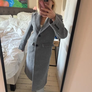 Grå lång kappa från H&M - Säljer en stilren grå kappa från H&M i klassisk modell med krage, två knappar framtill och fickor med lock. Lång passform och fodrad insida, perfekt för kyliga dagar. Snygg och enkel att matcha med allt.