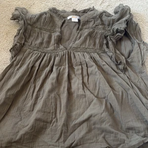 Grå blus med spets från Gina Tricot - Säljer en grå blus från Gina Tricot i storlek 146/152. Blusen har volangdetaljer vid axlarna, spetsband över bröstet och är tillverkad i ett mjukt, tunt bomullstyg. V-ringad hals och korta ärmar med rynkade detaljer ger en chill och feminin vibe.