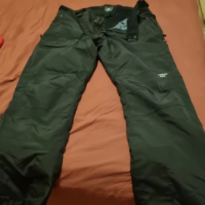 Svarta täckbyxor från North End - Svarta täckbyxor från North End med praktiska fickor och justerbar midja. Perfekta för kalla dagar och vinteraktiviteter. Byxorna har en klassisk passform och är tillverkade i ett slitstarkt syntetmaterial som står emot väder och vind.