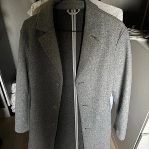 Grå kappa med klassisk krage - Snygg grå kappa med klassisk krage och tre knappar framtill. Rak passform och stilren design som passar perfekt till höst och vinter. Materialet känns mjukt och har en ull-liknande struktur. Långärmad och enkel att matcha med olika outfits.