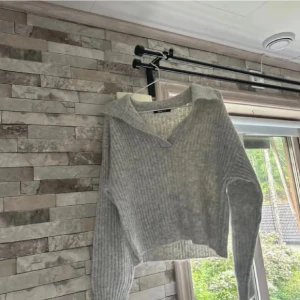 Grå ribbad tröja med krage - Säljer en grå ribbad croppad tröja med krage och v-ringning. Tröjan har lång ärm och är stickad i ett mjukt material, perfekt för lager-på-lager. Enkel och stilren design som passar till mycket.