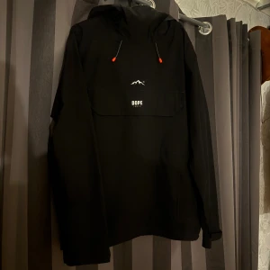Svart vindjacka från Dope - Svart vindjacka från Dope med huva och dragsko med röda detaljer. Stor ficka framtill med Dope-logga. Perfekt för dig som gillar streetwear och vill ha en stilren jacka med snygga detaljer. Endast använd fåtal gånger💞Säljer även 2 till dope jackor. Storlek M