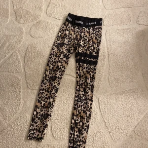 Leopardmönstrade leggings från Stronger - Snygga leopardmönstrade leggings från Stronger med bred svart resår i midjan där loggan är tryckt runtom. Tight passform och stretchigt material som passar perfekt till träning. Färgerna går i beige, brun och svart.