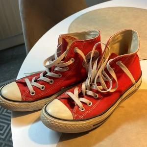 Röda Converse All Star high tops - Klassiska röda Converse All Star high tops med vit gummitå och sula. Skorna har vita snören, silverfärgade öljetter och tydlig logga på hälen. Tillverkade i textil med kontrastsömmar och platt sula. Perfekta för en cool och avslappnad stil.