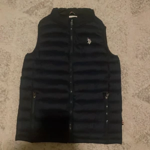 Mörkblå dunväst från U.S. Polo Assn. - Snygg Mörkblå dunväst från U.S. Polo Assn. med horisontella quiltade sömmar och dragkedja framtill. Västen har två fickor med dragkedja och en liten broderad logga på bröstet. Perfekt för lager-på-lager och stilren look. 