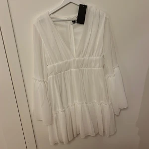 Vit omlottklänning med volanger - Superfin vit kortklänning med v-ringning och omlottkänsla. Klänningen har långa, vida ärmar och flera lager volanger som ger en luftig och romantisk look. Perfekt som studentklänning 