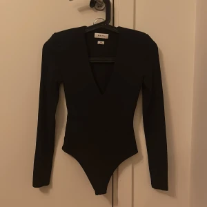 Svart långärmad topp från Babaton - Säljer en svart långärmad topp från Babaton med djup v-ringning och figurnära passform. Toppen är stilren och har en body-modell som sitter snyggt till höga jeans eller kjol. Perfekt för dig som gillar minimalistisk och trendig stil. Har shoulder pads och ger en elegant stilren ram.