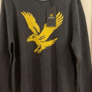 Grå stickad tröja Lyle & Scott - Mörkgrå stickad tröja från Lyle & Scott med stor gul örn på bröstet. Tröjan har rund hals med ljusgrå ribbad kant och långa ärmar. Perfekt för dig som vill ha en snygg och stilren look med en cool detalj framtill.
