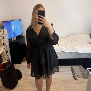 Svart omlottklänning med glitter - Supersnygg svart kort omlottklänning med diskret glittrigt tyg och långa ärmar. Klänningen har resår i midjan för en skön passform och volangdetalj i kjolen. Perfekt för dig som vill sticka ut lite extra!