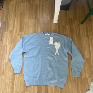 Ljusblå stickad tröja från Ami Paris - Säljer en ljusblå stickad tröja från Ami Alexandre Mattiussi med ett stort vitt hjärta och bokstaven A på bröstet. Tröjan har rund hals, ribbade muddar och är tillverkad i ekologisk bomull. Perfekt för dig som gillar stilrena och trendiga plagg.