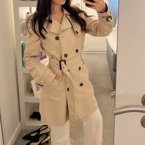 Beige trenchcoat - Stilren beige trenchcoat med dubbelknäppning, bälte i midjan och klassiska axelklaffar. Jackan har långa ärmar med spännen vid handlederna och snygga detaljer som stora knappar och fickor. Sååå snygg!!