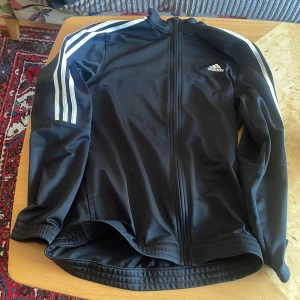 Svart Adidas vindjacka med vita ränder - Svart vindjacka från Adidas med klassiska vita ränder längs ärmarna och liten Adidas-logga på bröstet. Jackan har hel dragkedja framtill, hög krage och är tillverkad i ett glansigt syntetmaterial. Perfekt för en sportig look.