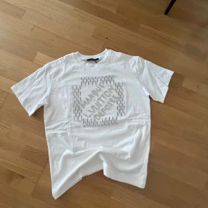 Vit Louis Vuitton t-shirt med pärldetaljer - Exklusiv vit t-shirt från Louis Vuitton med korta ärmar. Framsidan har ett broderat mönster och text i pärlor som ger en lyxig känsla. Tillverkad i Italien i mjuk bomull. Perfekt för dig som vill sticka ut med designerplagg och coola detaljer.