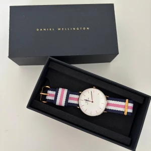 Daniel Wellington klocka med randigt band - Stilren klocka från Daniel Wellington med vitt ur och guldfärgade detaljer. Bandet är randigt i marinblått, rosa och vitt, vilket ger en fräsch och ungdomlig look. Klockan levereras i originalask.