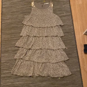 Leopardmönstrad volangklänning KappAhl - Supersnygg ärmlös klänning från KappAhl i beige med leopardmönster och flera volanglager. Axelbanden är dekorerade med glittriga paljetter som ger extra bling. Perfekt för dig som vill sticka ut med en cool och trendig look.