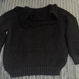 Svart stickad tröja offshoulder - Mysig svart stickad tröja med bred krage och långa ärmar. Tröjan har en chunky struktur och ribbade muddar vid ärmslut och nederkant. Perfekt för kyliga dagar och enkel att matcha med jeans eller kjol.