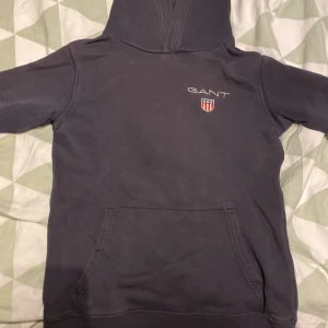 Mörkblå hoodie från GANT - Säljer en mörkblå hoodie från GANT med broderad logga och sköld på bröstet. Tröjan har huva, magficka och långa ärmar med ribbade muddar. Perfekt för dig som gillar klassisk och stilren streetwear.