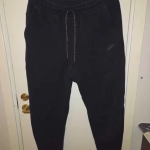 Svarta Nike tech byxor  - Svarta mjukisbyxor från Nike med dragsko i midjan och diskret logga på benet. Byxorna har en avslappnad passform och är tillverkade i mjukt bomullsmaterial. Perfekta för chill eller träning. Använda en gång dom är som helt nya 