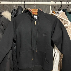 Svart hoodie från Burberry med dragkedja - Svart hoodie från Burberry med klassisk rutmönstrad insida i huvan. Tröjan har guldfärgad dragkedja, snörning vid huvan och två stora fickor framtill. Materialet är mjuk bomull och passformen är normal. Perfekt för dig som gillar stilrena märkesplagg.