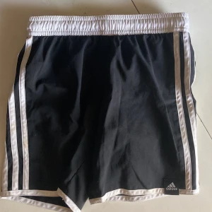 Svarta Adidas shorts med vita ränder - Snygga svarta Adidas shorts med klassiska vita ränder längs sidorna och vit resår i midjan. Shortsen har meshfoder på insidan och är perfekta för sport eller chill. Adidas-logga framtill och en bakficka för småsaker.