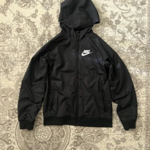 Nike - Svart vindjacka med huva - Svart vindjacka från Nike med vit logga på bröstet. Jackan har huva, dragkedja framtill och två fickor med dragkedja. Tillverkad i lätt polyester med meshfoder för extra komfort. Perfekt för blåsiga dagar och sportiga outfits. Barnstorlek L men passar som S/M för tonåringar/vuxna. Inga tecken på använding.