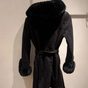 Svart fuskpälsjacka med bälte från Giorgia fashion - Snygg svart fuskpälsjacka från Giorgia fashion med stora fluffiga pälskanter vid krage med luva och fluffiga ärmslut. Jackan har bälte i midjan och dragkedja på fickorna. Materialet är mjukt och matt , med en lyxig känsla. Perfekt för dig som vill sticka ut med en statementjacka i vinter.