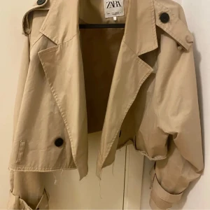 Beige croppad trenchcoat från Zara - Snygg croppad trenchcoat från Zara i beige bomullsmaterial. Jackan har stora svarta knappar, axelklaffar och bälte vid ärmsluten. Rå kant nertill för en trendig look och oversized passform. Perfekt för dig som vill sticka ut med en modern twist på klassikern. Sitter dock som M/S