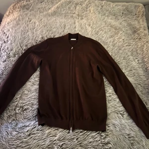 Brun kofta från John Henric, M - Snygg brun kofta från John Henric i storlek M. Koftan har hel dragkedja framtill, ribbade muddar och är tillverkad i ett mjukt, stickat material. Perfekt för lager-på-lager och funkar året runt. Enkel och stilren design som passar till det mesta.